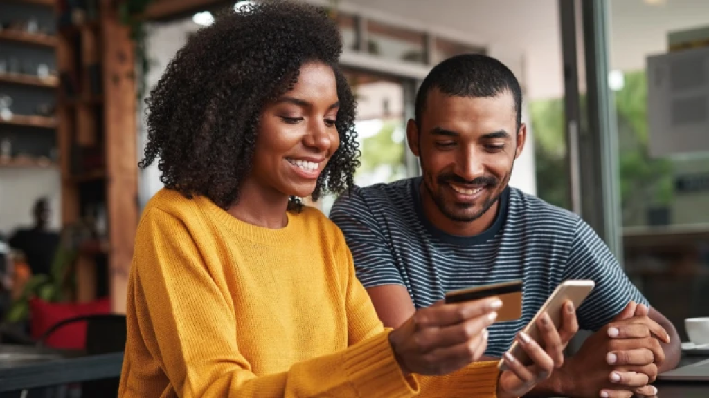 Two people sitting next to each other smiling. One person is holding a credit card in one hand and a smartphone in the other.